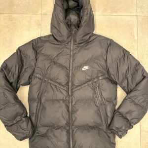Svart pufferjacka från Nike - Svart pufferjacka från Nike med huva och dragkedja framtill. Jackan har två snedställda fickor med dragkedja och diskret Nike-logga på bröstet.
