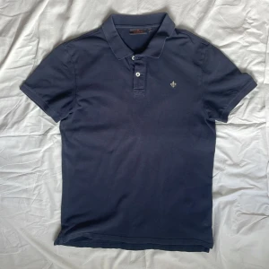 Mörkblå pikétröja från Morris - Snygg mörkblå pikétröja från Morris med klassisk krage och två knappar framtill. Tröjan har korta ärmar och är tillverkad i bomull. På bröstet finns en broderad logga i vitt. Perfekt för en clean och stilren look.