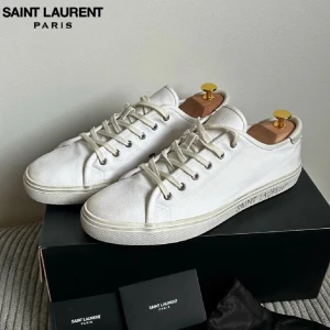 Vita Saint Laurent sneakers i sjukt bra skick (10/10). Nypris på ca 8000kr  - Snygga vita sneakers från Saint Laurent Paris i canvas med klassisk låg profil och rund tå. Skorna har snörning med metallöljetter och diskret logga på sidan av sulan. Perfekta för en clean och stilren look.