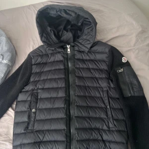 Svart Moncler pufferjacka med luva - Säljer en svart pufferjacka från Moncler med stickade ärmar och detaljer. Jackan har huva, tvåvägsdragkedja och ribbad struktur på ärmar och rygg. Märkespatch på ena ärmen och praktiska fickor framtill. Perfekt för dig som vill ha en snygg och varm jacka med streetkänsla.