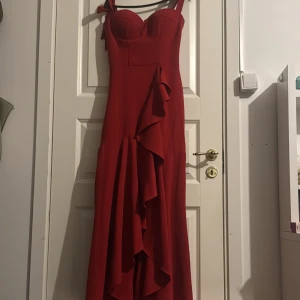 Röd långklänning med  band som kan knyta  - Säljer en elegant röd långklänning från MK Mode med axelband som går att knyta. Klänningen har en snygg v-ringad byst, volangdetaljer framtill och figurnära passform. Perfekt för bal eller fest! Materialet är mjukt och följsamt polyester.  Original pris var 1700kr