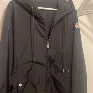 Svart vindjacka från Moncler - Snygg svart vindjacka från Moncler med huva och dragkedja framtill. Jackan har silverfärgade knappar med Moncler-logga, justerbar snörning vid huvan och diskret logotyp på ärmen. Materialet är lätt och syntetiskt, perfekt för blåsiga dagar. Fjärde bilden är enda defekten på jackan. NFC finns och QR funkar.