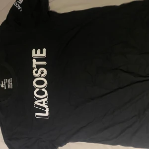 Svart Lacoste t-shirt - Svart t-shirt från Lacoste med stort vitt logotryck på bröstet och mindre logga med krokodil på ärmen. Rund hals och klassisk passform. Tillverkad i mjuk bomull som är skön mot huden. Perfekt för dig som gillar stilrena och sportiga märkesplagg. Bra skick, pris ka diskuteras!