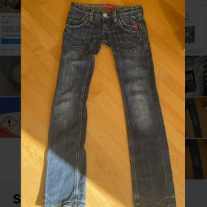 Blå jeans från My God med raka ben - Säljer ett par blå jeans från My God med snygg slitning och raka ben. Jeansen har klassisk femficksdesign, kontrastsömmar och en röd detalj på fickan. I storlek 27w34l. Bak finns en stor patch med röd text och grafik. Perfekta för en avslappnad streetstil.