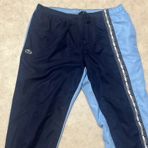 Lacoste Sport blå träningsbyxor L - Snygga träningsbyxor från Lacoste Sport i två nyanser av blått med logoband längs benet och klassisk krokodilpatch. Byxorna har elastisk midja med snörning och är tillverkade i ett lätt syntetmaterial. Perfekta för sportiga dagar eller chill.