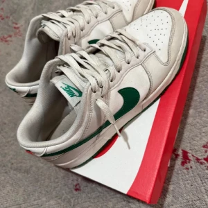 Nike Dunk Low vit/grön - Nike Dunk Low sneakers i vitt skinn med gröna detaljer och klassisk grön swoosh. Lågt skaft, perforerad tåbox och vit sula med grön yttersula. Perfekta för dig som gillar stilrena och sportiga sneakers med retrovibbar. Behöver bara tvättas.