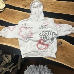 Vit hoodie med graffiti från Bershka - Vit hoodie från Bershka med unika graffiti-inspirerade tryck i rosa och svart. Hoodien har luva, långa ärmar och coola detaljer som text, stjärnor och blommor. Perfekt för dig som vill sticka ut med en streetstyle-look.