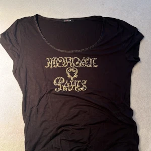 Svart t-shirt Morgan Paris tryck - Svart t-shirt från Morgan med coolt guldfärgat 'Morgan Paris'-tryck framtill i snirklig stil. T-shirten har rund halsringning och korta ärmar. Skön och lätt i materialet, perfekt till jeans för en chill look. Passar både M och S, jag på bilden har M