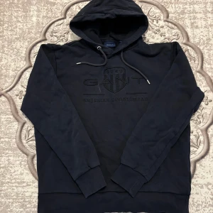 Svart hoodie från GANT med brodyr - Svart hoodie från GANT med broderad logga och text på bröstet. Klassisk modell med huva och dragsko, ribbade muddar och mjukt material. Perfekt för en chill och stilren look. Priset är inte i hugget’ så kan komma överens om något! den har tappat lite färg så därför de ser ut så på bilden. Den är mörk blå 