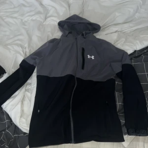 Grå och svart vindjacka från Under Armour - Snygg tvåfärgad vindjacka från Under Armour i grått och svart. Jackan har huva med dragsko, hel dragkedja framtill och praktiska fickor. Perfekt för dig som gillar sportig stil och vill ha något lätt och funktionellt.