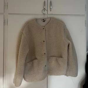 Beige teddyjacka från Divided M - Mysig beige teddyjacka från Divided i storlek M. Jackan har rund hals, knäppning framtill och stora fickor. Perfekt för kalla dagar när du vill vara både varm och snygg. Oversized passform och långa ärmar för en chill vibe.