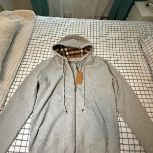 Grå Burberry hoodie med rutigt foder - Snygg grå hoodie från Burberry med dragkedja och klassiskt rutigt foder i huvan. Tröjan har långa ärmar, snörning i huvan och två fickor framtill. Perfekt för dig som vill ha en stilren men ändå lyxig look.