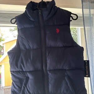 Mörkblå dunväst från U.S. Polo Assn. - Snygg mörkblå dunväst från U.S. Polo Assn. med hög krage och dragkedja framtill. Västen har två fickor och en röd broderad logga på bröstet. Perfekt för lager-på-lager och chill höst- eller vinterstil. Aldrig använd, pris kan diskuteras.