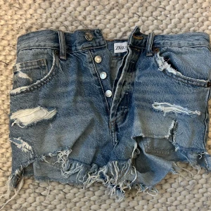 Blå slitna jeansshorts från Zara - Snygga blå jeansshorts från Zara, slitna detaljer och fransig kant. Klassisk femficksmodell med knappar framtill. Perfekta för sommardagar när du vill ha en avslappnad och trendig look.