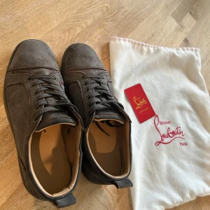 Grå mocka sneakers från Louboutin - Snygga grå sneakers från Christian Louboutin i mjuk mocka med klassisk snörning och rund tå. Yttersulan är röd med Louboutin-logga, vilket ger en unik och lyxig touch. Perfekt för dig som gillar exklusiva detaljer och stilren design. Priser ej hugget i sten, hör av dig vid funderingar!
