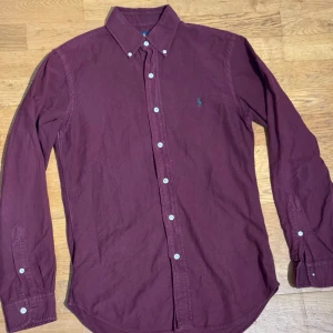 Vinröd skjorta från Ralph Lauren - Slim fit vinröd skjorta från Ralph Lauren med klassisk krage och knappar hela vägen. Broderad logga i grönt på bröstet. Långärmad och tillverkad i mjuk bomull som känns skön mot huden. Perfekt för dig som gillar stilrena plagg med lite edge. En knapp på vänster arm har lossnat med extra knappar finns i skjortan 
