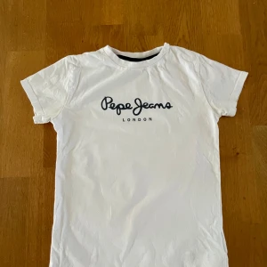 Vit t-shirt Pepe Jeans London - Vit t-shirt från Pepe Jeans London med svart logotyptryck på bröstet. Klassisk rund hals och korta ärmar. T-shirten har en clean och enkel design, perfekt för dig som gillar stilrena plagg med snygg branding. Materialet är mjuk bomull som känns skönt mot huden.