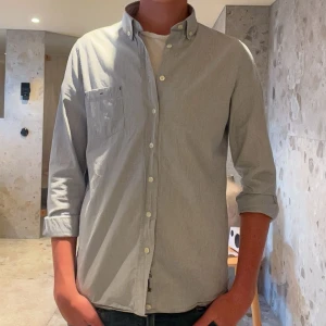 Ljusblå randig skjorta med knappar - Snygg ljusblå skjorta med tunna vita ränder och klassisk button-down krage. Skjortan har långärmade ärmar, bröstficka och vita knappar framtill. Tillverkad i mjuk bomull som känns skön mot huden. Perfekt för dig som gillar stilrena och fräscha plagg.