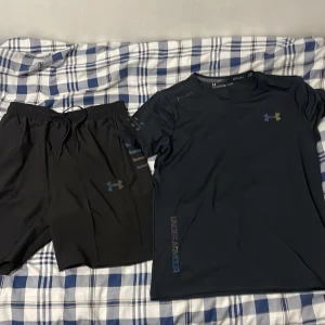 Svart Under Armour HeatGear  - Svart Under Armour t-shirt i tekniskt HeatGear-material, perfekt för träning. T-shirten har korta ärmar, rund hals och en diskret logga i regnbågston på bröstet samt Under Armour-tryck längs nederkanten. Slimmad passform som sitter snyggt och bekvämt.