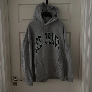 Grå hoodie från Lee Jeans - Snygg grå hoodie från Lee Jeans med stor logga i grönt och vitt på bröstet. Tröjan har huva med snörning och en stor magficka. Perfekt för en avslappnad och cool stil.