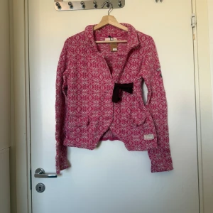 Mönstrad rosa cardigan jacka Odd Molly - Superfin rosa  kofta från Odd Molly med vitt mönster och snygg rosett i sammet framtill. Kofta är stickad i mjukt material, har lång ärm och två fickor framtill. Perfekt för dig som gillar färg och unika detaljer! Storlek 2 som enligt deras storleksguide motsvarar en storlek M. Mycket fint skick, men ytludd och noppror kan förekomma eftersom materialet är ull och nylon. 