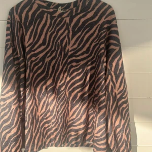 Zebramönstrad tröja från Nelly.com - Säljer en snygg långärmad tröja från Nelly.com i storlek M. Tröjan har ett coolt zebramönster i svart och beige, perfekt för dig som vill sticka ut. Mjuk och skön med rund halsringning, passar till både jeans och kjol.