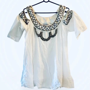 Vit tunika med paljettdetaljer Fashionmob - Supersnygg vit tunika från Fashionmob med unika paljettdetaljer och pärlor i silver och svart runt halsringningen och axlarna. Blusen har korta ärmar med cut out vid axlarna och en lös passform. Perfekt för dig som vill sticka ut med coola detaljer. 