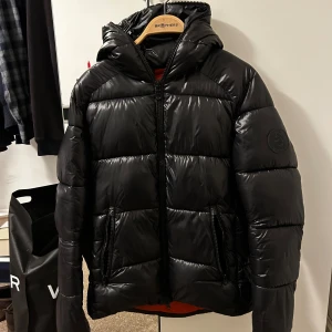 Svart pufferjacka från Save The Duck - Säljer en svart pufferjacka från Save The Duck med glansig finish och orange foder. Jackan har huva, två dragkedjefickor och hel dragkedja framtill. 100% animal free och perfekt för kalla dagar. Snygg och stilren design som sticker ut med den orangea insidan.