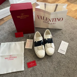 Valentino Garavani Open sneakers vit/blå - Snygga Valentino Garavani Open sneakers i vitt skinn med bred mörkblå rem över vristen. Klassisk rund tå, platt sula och coola nitar på hälen. Guldlogga på plösen och vita snören. Kommer med originalkartong, dustbag och äkthetskort.