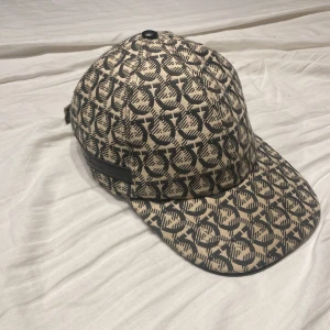 Beige och svart keps från Salvatore Ferragamo - Snygg keps från Salvatore Ferragamo i beige och svart med ikoniskt mönster över hela kepsen. Justerbar rem baktill med metallspänne. Tillverkad i bomullsmaterial och har en klassisk böjd skärm. Perfekt för dig som vill sticka ut med en exklusiv accessoar. Pris går att diskutera och byten kan vara intressant. Kepsen har gått igenom en äkthetskontroll och kan skicka kvitto ifrån den om du vill.