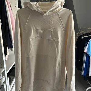 Vit hoodie från J.Lindeberg, M - Säljer en stilren hoodie från J.Lindeberg i färgen cloud white. Tröjan har en stor huva, broderad logga framtill och en rymlig magficka. Tillverkad i mjuk bomull och har en relaxed passform med långa ärmar. Perfekt för en clean och modern look. Nypris 2399