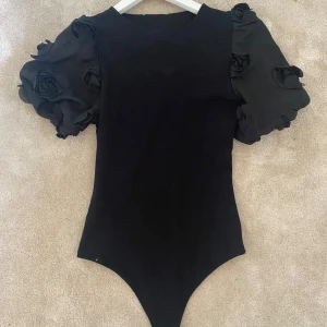 Svart body med puffärmar och rosdetaljer - Supersnygg svart body med stora puffärmar i tyg och 3D-rosdetaljer. Kroppsnära passform och rund halsringning. Ärmarna är korta och ger en riktigt cool statement-look. Perfekt för dig som vill sticka ut med en unik topp!