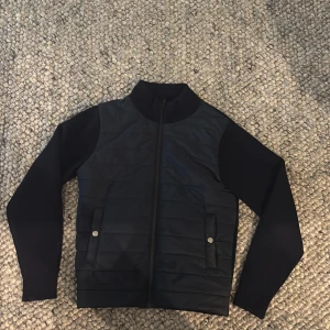 Marinblå cardiganjacka från Sayless - Marinblå cardiganjacka i ull från Sayless med quiltad framsida, två fickor med tryckknappar och hel dragkedja. Jackan har långärmade släta ärmar och ribbad krage. Perfekt för dig som gillar stilren och enkel design. Storlek S. 