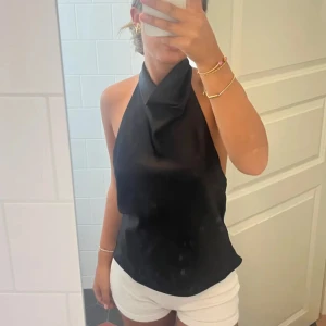Svart halterneck topp med öppen rygg - Säljer en svart halterneck topp med snygg drapering framtill och helt öppen rygg. Toppen har en elegant och stilren look, perfekt för dig som vill sticka ut. Materialet känns slätt och svalt mot huden. Passar dig som gillar minimalistisk och trendig stil.