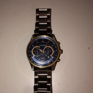 Citizen herrklocka i silver och guld - Stilren Citizen herrklocka med blå urtavla, silverfärgat armband i metall och snygga gulddetaljer. Klockan har kronograf, datumvisning och tachymeter runt urtavlan. Länkarna är blanka med guldfärgade linjer för en extra lyxig känsla.