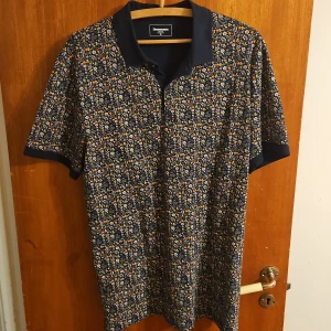 Blommig pikétröja från Dressmann XL - Snygg pikétröja från Dressmann i storlek XL med mörkblå krage och ärmslut. T-shirten har ett blommigt mönster i vitt, gult och orange på marinblå botten. Perfekt för dig som vill sticka ut med en färgglad och unik stil.