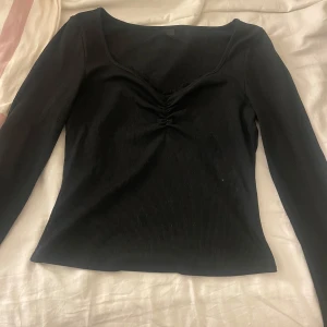 Svart ribbad långärmad topp - En svart långärmad topp med ribbad struktur och snygg rynkdetalj vid bysten. Djup, rundad ringning framtill.