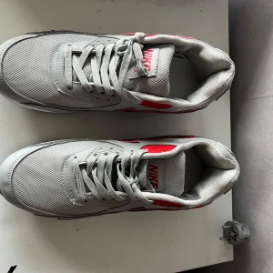 Nike Air Max 90 grå/röd sneakers - Snygga Nike Air Max 90 sneakers i grått med svarta och röda detaljer. Skorna har mesh och syntet på ovandelen, klassisk Air Max-sula och snörning. Perfekta för dig som gillar streetstyle och vill ha en ikonisk sneaker med cool färgkombo.