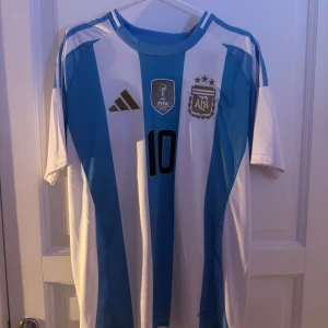 Argentina Messi 10 Adidas fotbollströja L - Säljer en officiell Argentina fotbollströja från Adidas i storlek L, med Messis namn och nummer 10 på ryggen. Tröjan har klassiska blå och vita ränder, korta ärmar och broderade märken på bröstet. Slim fit-modell i lätt och ventilerande material.