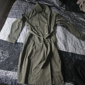 Olivgrön trenchcoat från SHEIN - Olivgrön trenchcoat från SHEIN med klassisk krage, knappar framtill och matchande bälte i midjan. Den har långa ärmar med knappdetalj vid ärmslut och är tillverkad i bomull. Perfekt för dig som gillar stilrena och tidlösa plagg.
