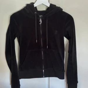 Svart velour hoodie Juicy Couture XXS - Svart hoodie i mjuk velour från Juicy Couture. Tröjan har huva, snörning, dragkedja med JC-logga och känguruficka framtill. Perfekt för en chill och trendig look. Materialet är 95% polyester och 5% elastan för extra stretch och komfort.