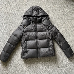 Svart pufferjacka från Calvin Klein - Säljer en svart pufferjacka från Calvin Klein med huva och dragkedja framtill. Jackan har två fickor med dragkedja och diskret CK-logga på bröstet. Perfekt för kalla dagar och riktigt snygg till streetwear-stilen.
