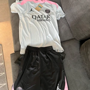 PSG x Jordan fotbolls set  ljusgrå/rosa - Snygg PSG x Jordan fotbollströja i ljusgrått med rosa detaljer på axlarna och svart logga. Tröjan har korta ärmar och är gjord i ett lätt, sportigt material som andas. Paris Saint-Germain-emblem och Qatar Airways-tryck på bröstet. Perfekt för dig som gillar streetwear och fotboll. Även shorts med fickor och dragkedja 