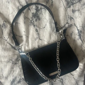 Svart axelväska med kedja från URBAN OUTFITERS - Snygg svart axelväska med croco-mönstrad yta och en silverfärgad kedja som detalj. Väskan har en dragkedja upptill och en metallbricka med logga på framsidan. Perfekt storlek för det viktigaste och ger en cool vibe till din outfit. 