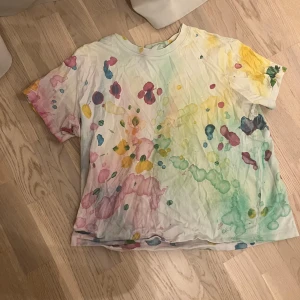 Färgstark t-shirt från H&M i M - Unik t-shirt från H&M i storlek M med färgglada splash-mönster i rosa, gul, grön och blå på vit botten. Modellen är klassisk med rund hals och korta ärmar. Materialet är mjuk bomull, perfekt för dig som gillar kreativa och färgstarka plagg.