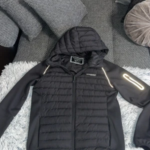 Svart pufferjacka från McKenzie - Svart pufferjacka från McKenzie med huva och dragkedja framtill. Jackan har quiltad framsida och rygg, släta ärmar med reflekterande detalj på ena ärmen och en sportig look. Perfekt för kyliga dagar när du vill hålla stilen enkel och clean. PRIS KAN DISKUTERAS!