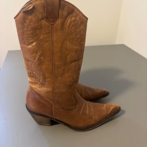 Bruna cowboy boots med spetsig tå - Säljer ett par snygga bruna cowboy boots i skinn med spetsig tå och låg klack. Bootsens skaft har broderade fjärilsmönster som ger dem en unik vibe. Perfekta till jeans eller klänning för en cool westernstil.