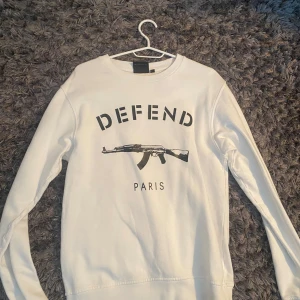 Vit Defend Paris sweatshirt - Säljer en vit sweatshirt från Defend Paris med svart tryck framtill. Tröjan har rund hals, långa ärmar och ett stort grafiskt motiv med texten 'DEFEND PARIS' och en AK-47. Perfekt för dig som gillar streetwear och statement-plagg.