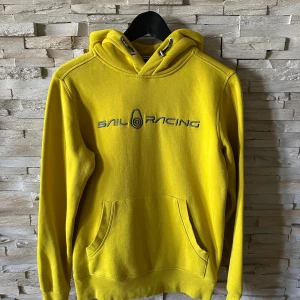 Gul hoodie från Sail Racing - Gul hoodie från Sail Racing med broderad logga på bröstet och en mindre logga på huvan. Tröjan har känguruficka, snörning i huvan och långa ärmar. Perfekt för dig som gillar sportig och färgstark stil.