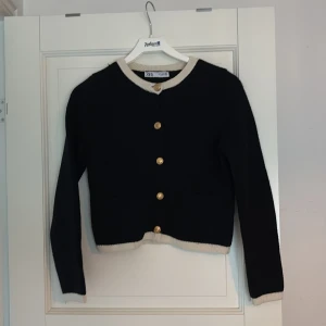 Svart kofta med guldknappar från Zara - Stilren svart kofta från Zara med långa ärmar, guldiga knappar och kontrasterande beige kant vid hals, ärmslut och nederkant. Croppad modell/midjekort